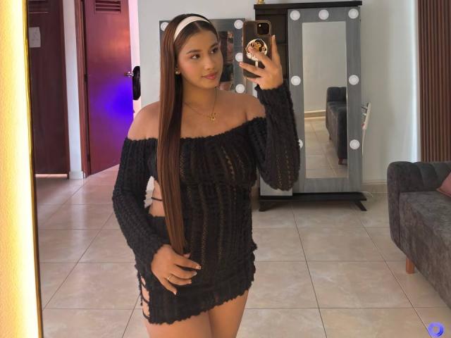SofiaSweetty mit langen braunen Haaren, trägt ein schwarzes, trägerloses Strickkleid und macht ein Selfie vor einem Spiegel mit ihrem Handy. Dabei zeigt sie ihr Gesäß und ihre kleinen Brüste, und das Ganze spielt sich in einem Raum ab..