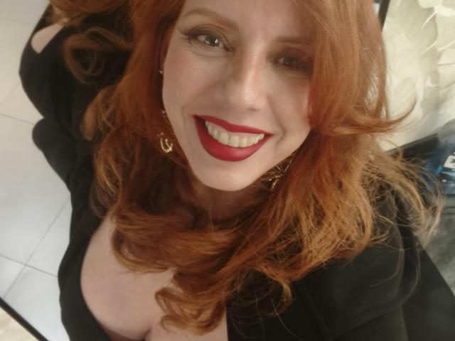 Khharha ha i capelli rossi, indossa una maglia nera, sorride con il rossetto rosso, mostra il décolleté e porta orecchini dorati. Sta guardando verso l'alto, scattando un selfie..