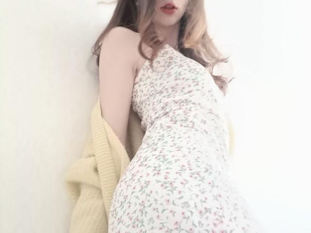 WollyMolly with long brown hair, red lips, floral dress, yellow cardigan, leaning against white wall, soft light, showing her small tits and curvy ass.