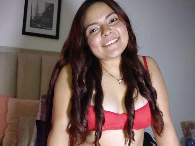 CherryScarlett, a curvy woman with long red wavy hair, wearing a red bra, smiling with shiny skin, showing her tits. She's in a bedroom with a beige headboard.