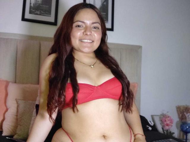 CherryScarlett with long red hair, smiling, wearing a red bra and thong, showing her tits and a small belly, standing in a bedroom.