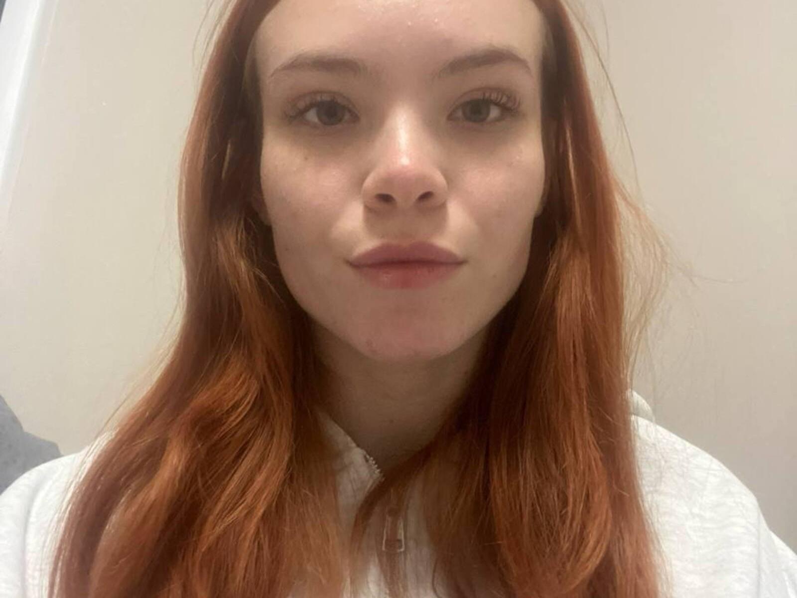 SimonaZeiler has red hair, fair skin, and a neutral expression. She's wearing a white shirt with a buttoned collar. Her nose is centered, and she's looking directly at the camera.