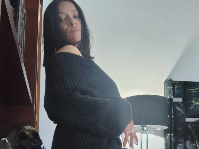 Amelie Blake mit schulterlangem, schwarzem Haar, trägt einen schwarzen Strickpullover mit einem tiefen Ausschnitt und steht seitlich. Sie hat ein kleines Nasenpiercing. Im Hintergrund sind eine schwarze Lampe und ein Regal zu sehen..