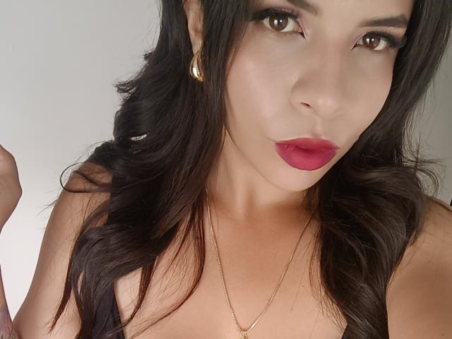 VioletaStone, avec ses cheveux noirs et ondulés, ses yeux marrons, son rouge à lèvres rose, ses boucles d'or et son haut noir, dévoilant légèrement sa poitrine. Elle porte un collier en or..