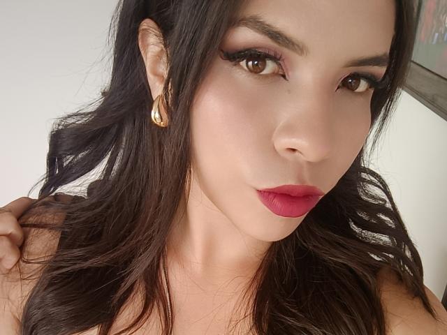 VioletaStone hat dunkles, welliges Haar, braune Augen, roten Lippenstift und goldene Ohrringe. Sie schaut in die Kamera, ihre rosa Lippen sind leicht geöffnet, und sie hat eine Hand in ihrem Haar..