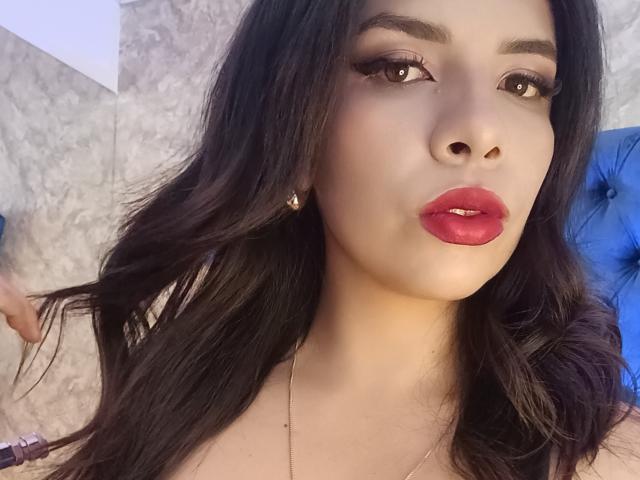 VioletaStone, avec ses longs cheveux noirs, son rouge à lèvres rouge et ses longs cils, portant de petites boucles d'oreilles, regarde l'appareil photo avec une légère moue, ses seins ne sont pas visibles sur la photo..