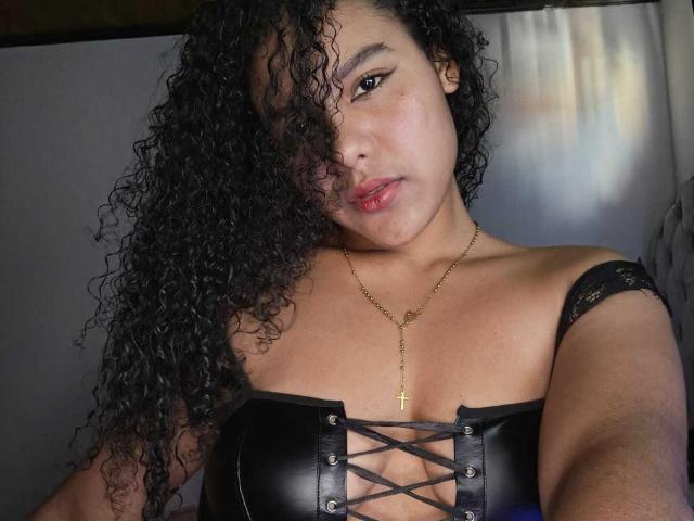 AriaVega with curly black hair, wearing a black leather corset, showing her tits, gold necklace, and pouty pink lips, taking a selfie, looking at the camera.