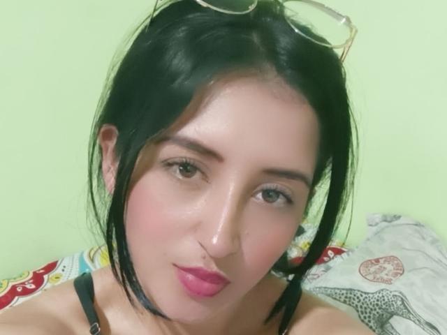 NicollJeans with black hair, pink lipstick, and glasses on head, showing off her nose and smiling slightly, wearing a black strap top, against a light green wall.