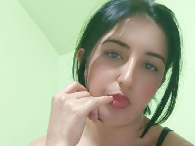NicollJeans has black hair, fair skin, and is pouting with her finger in her pink, glossy lips. She's wearing a black strap, and her eyes are half-closed.