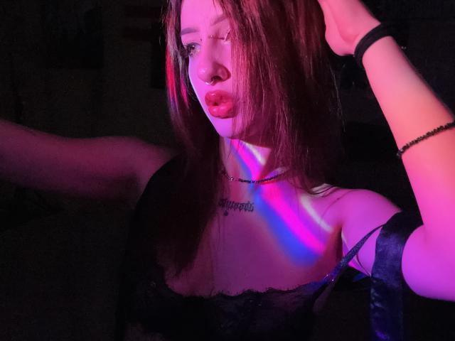 SimonneBrustkern with pink and purple neon lights on her face and chest, wearing black lace, raising her arm, has red lips, nose ring, and tattoo under her chin.