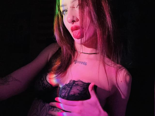 SimonneBrustkern with pink and green lighting on her face, red lipstick, black lace bra, hand covering her tits, tattoo on chest, choker necklace, long brown hair, dark background.