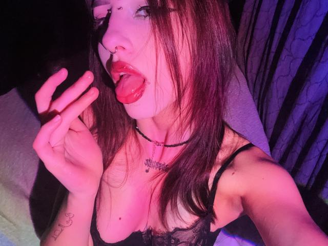 SimonneBrustkern with tongue out, licking fingers, wearing black lace bra, nose ring, choker, and tattoo on wrist, under pink-blue light, showing small tits and tattooed chest.