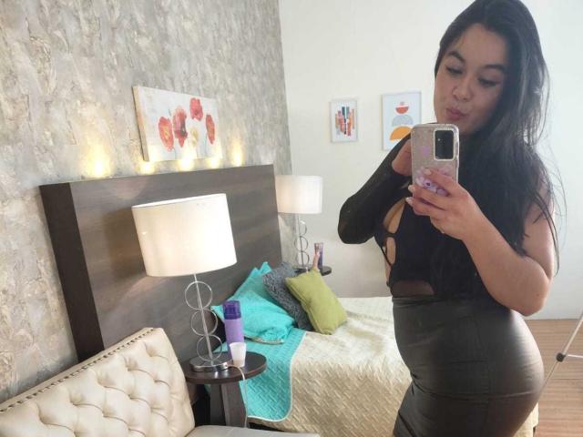 EveWillis with long black hair, wearing a black cut-out top and tight gray leather pants, taking a mirror selfie showing her curvy ass and small tits.