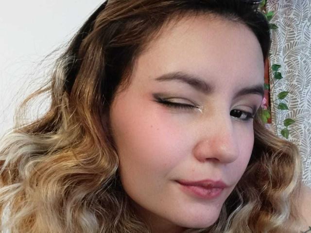 AlanisBlanc's winking face with curly blonde hair, light makeup, and pink lips. She's got a cute nose and slightly flushed skin. Her black eyeliner is noticeable.