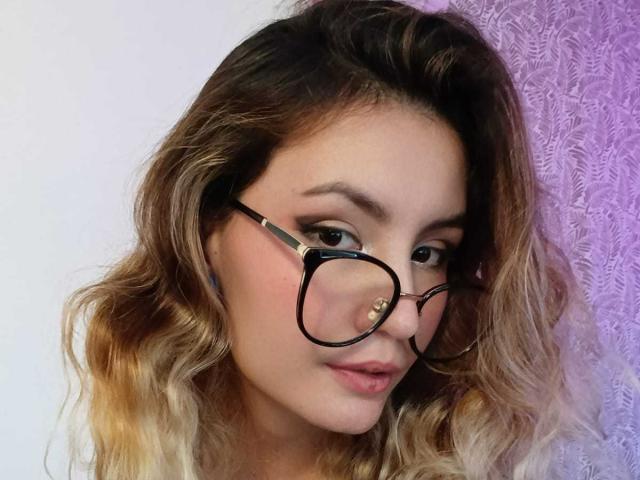 AlanisBlanc has wavy blonde hair, black-rimmed glasses, and a nose ring. Her makeup is subtle, with pinkish lips. She looks slightly down with a cute smile.