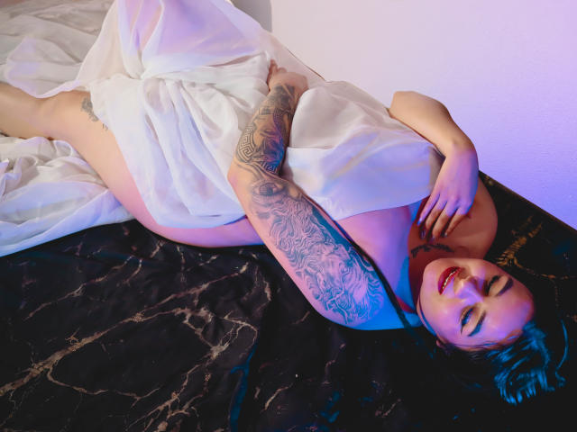 EveWillis lies on a black marble bed, covered in a white sheet, showing a tattooed arm and red lipstick, with a relaxed, sexy expression. Her pussy is partially visible.