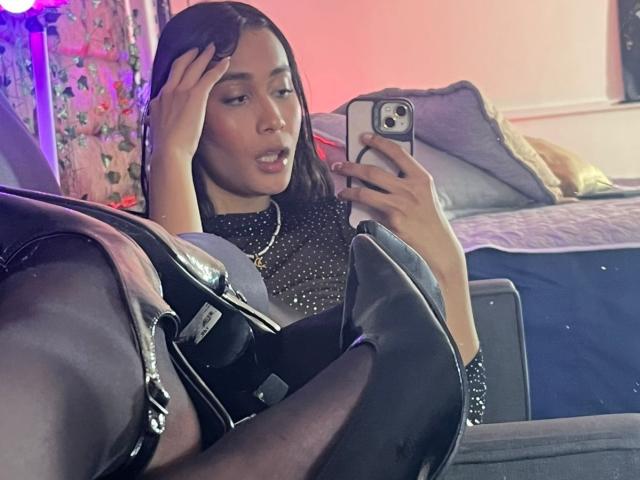 JennTylor, con i capelli scuri, indossa una maglia nera scintillante e sta scattando un selfie allo specchio, con una mano sulla testa. In primo piano si vedono degli stivali neri. Ha un'espressione leggermente confusa, e sullo sfondo ci sono luci rosa e viola..