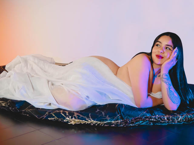 EveWillis está acostada en una cama negra, cubierta con una sábana blanca, con las nalgas levantadas y los pechos parcialmente visibles. Tiene el pelo largo y negro, tatuajes y una apariencia sexy..