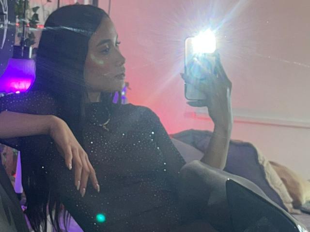 JennTylor, avec ses longs cheveux noirs, portant un haut noir scintillant, prend un selfie dans un miroir avec un flash puissant, penchée en arrière dans une chaise, le bras droit reposant..