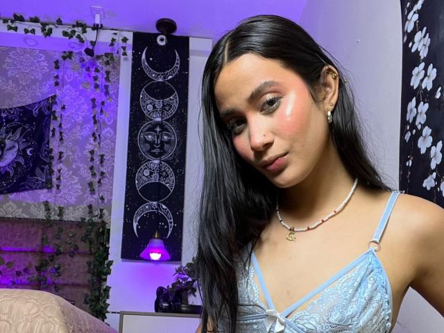 JennTylor, avec de longs cheveux noirs, portant un soutien-gorge en dentelle bleu clair, avec un blush rose sur les joues, un collier, des boucles d'oreilles et un sourire discret, dans une pièce décorée en violet et blanc..