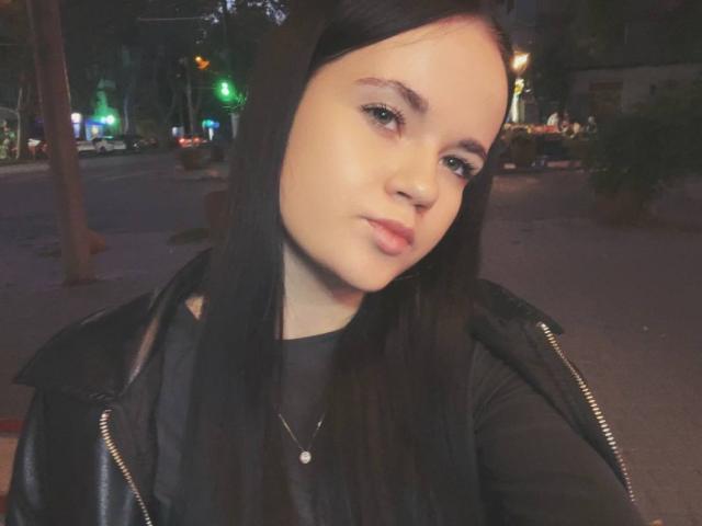 RoxyVesper has pale skin, straight black hair, and is pouting with pink lips. She's wearing a black leather jacket and a small necklace. Nighttime, city street background.