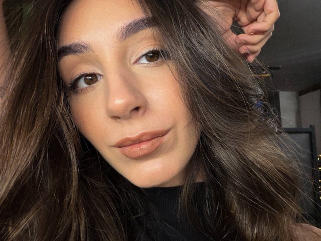 SmarteLiz has brown eyes, wavy brown hair, and full lips with a slight pout. She's wearing a black top, looking relaxed with a hand in her hair.