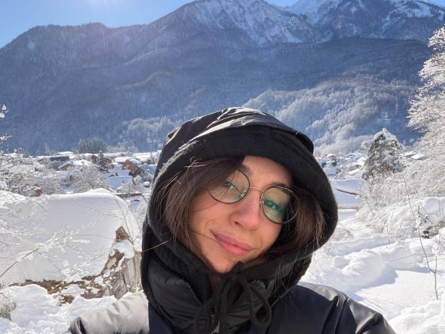 SmarteLiz with round glasses, black puffy jacket, and black hood, smiling in snowy landscape, mountains in background, brown hair peeking out, clear blue sky, reflective glasses.