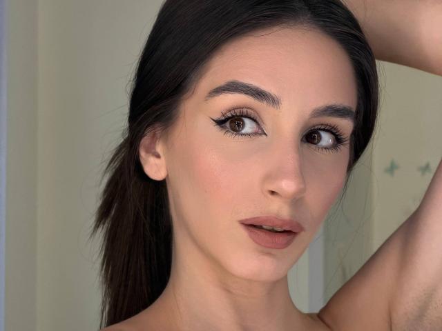 SmarteLiz has brown eyes, dark eyebrows, long eyelashes, and brown lipstick. She's looking at the camera with her arm raised, showing her armpit. Her skin is fair and smooth.
