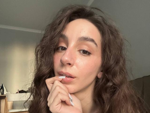 SmarteLiz, met krullend bruin haar, een lichtrode huid en een bedachtzame uitdrukking, bijt in haar witte nagel en kijkt naar de camera. Ze draagt subtiele make-up en een zwarte top..