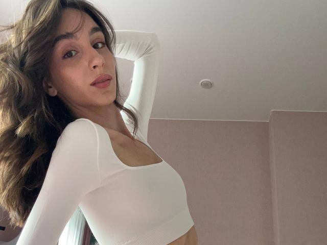 SmarteLiz mit langen, braunen, gewellten Haaren, trägt ein weißes, langärmeliges Crop-Top, das ihre kleinen Brüste zeigt, und hat eine schlanke Taille. Sie blickt mit leicht gerunzelter Miene in die Kamera..