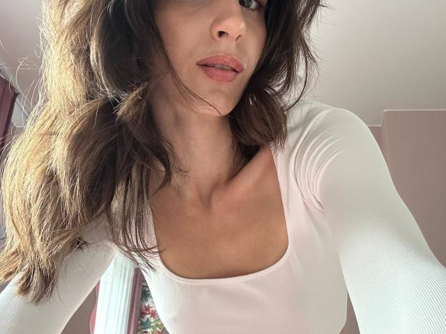 SmarteLiz with wavy brown hair, pink lips, and a white long-sleeve top, showing her small tits and slim neck in a low-angle selfie.