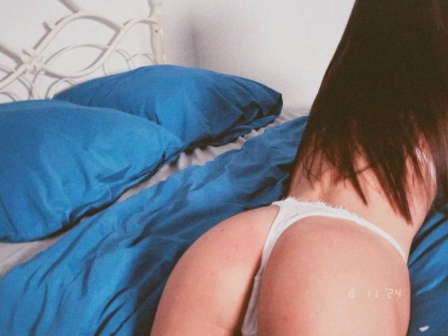 SmartLiz's ass is facing the camera, wearing white thong underwear, lying on a bed with blue pillows and white sheets. Her brown hair is down, covering part of her back.