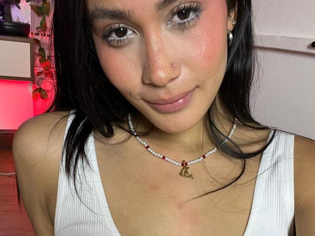 JennTylor with brown eyes, black hair, white tank top, pearl necklace, small nose ring, slight smile, pinkish skin, and small freckles on her nose. Boobs visible, small to medium size.
