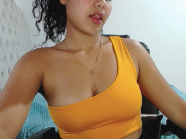 AriaVega, con i capelli neri ricci, indossa una maglietta gialla con una spalla scoperta che mette in mostra il seno, una collana d'oro e rossetto rosso, con un'espressione leggermente a bocca aperta. La sua pelle abbronzata contrasta con la maglietta gialla..
