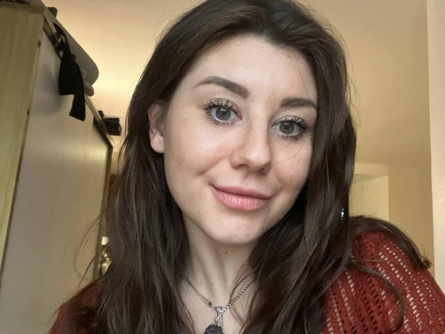 Cecily Axt, com cabelos castanhos longos, olhos azuis, lábios rosados e um leve sorriso, veste uma blusa de tricô vermelha, tem um pequeno piercing no nariz e um colar com um pingente..