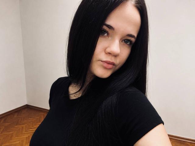 RoxyVesper has long black hair, blue eyes, and is wearing a black shirt, showing her small tits and a neutral expression in a simple room with wooden floors.