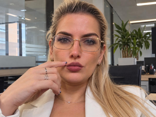 GiuliaAddams a les cheveux blonds, porte des lunettes et une chemise blanche, avec son doigt touchant ses lèvres charnues, elle porte une bague et des boucles d'oreilles, et se trouve dans un bureau avec une plante..