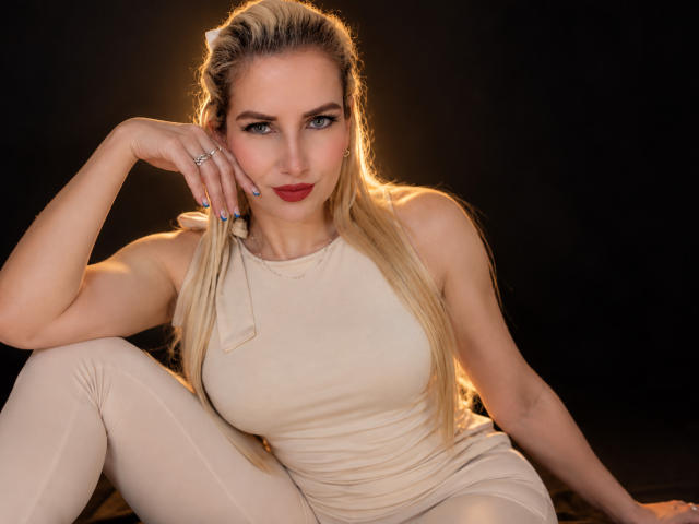 GiuliaAddams, con capelli biondi, rossetto rosso e unghie blu, indossa una maglia beige e pantaloni abbinati, seduta di fronte a uno sfondo scuro, con una mano sul viso, e guarda verso la fotocamera..