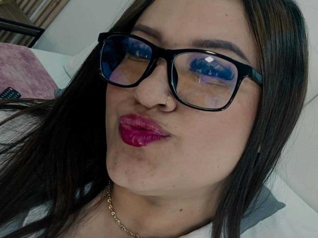 ValeriaaRose with pink lipstick, black glasses, nose ring, and gold chain, pouting her plump ass in a selfie. Her brown hair is straight, and she's lying down.