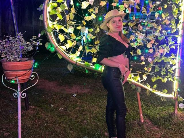ValeriaaRose with a tan hat, black tied-up shirt, and black leather pants, standing by a glowing vine-covered circular frame, looking up with a confident, sexy expression.