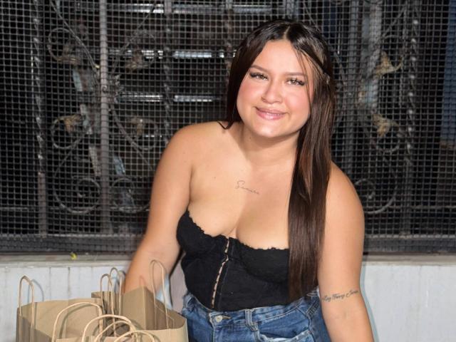 ValeriaaRose with medium tits, wearing a black strapless top and blue jeans, smiling, holding brown paper bags, has tattoos on chest and arm, long brown hair, in front of metal cage.