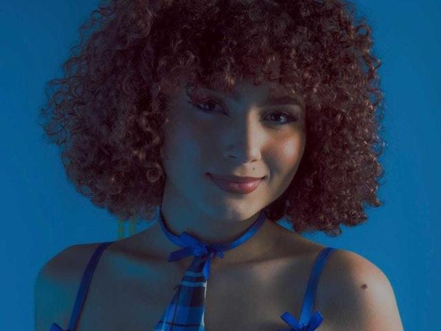 MakiFoxy con capelli castano ricci, reggiseno blu e cravatta abbinata, sorride leggermente, sfondo blu. Seno visibile, di medie dimensioni, viso carino, pelle color nocciola, piccola lentiggine sulla spalla destra..