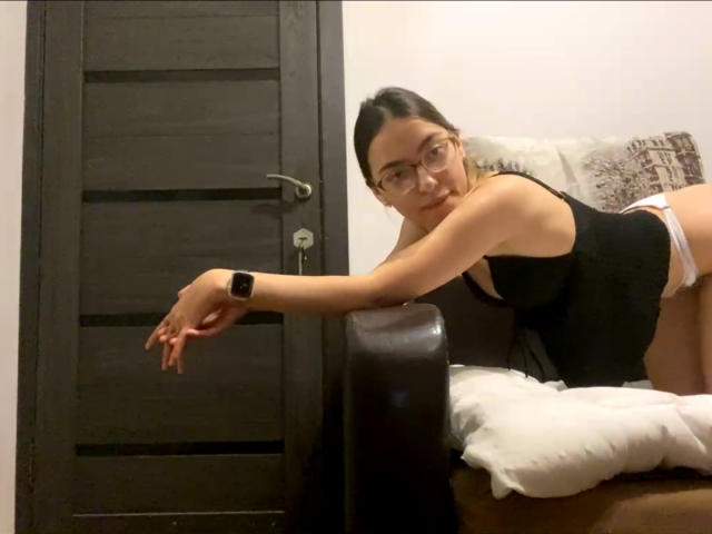 RaneeDerienzo with glasses, black tank top, white panties, leaning on brown armchair, black door in background, smartwatch on wrist, looking at camera, smiling.