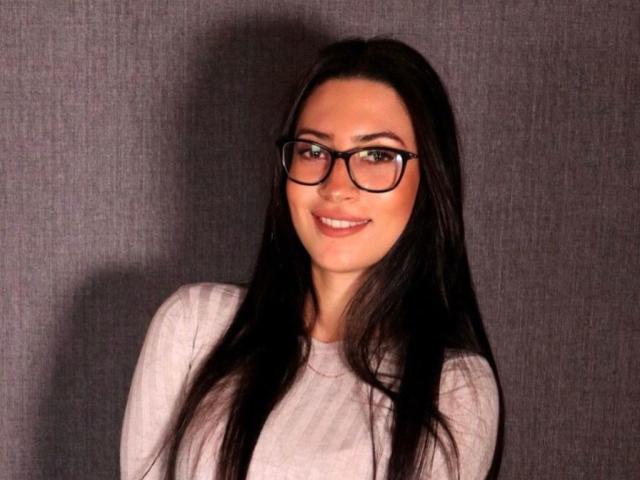 Victoriafs hat langes, schwarzes Haar, trägt eine schwarze Brille und einen hellrosa Pullover und lächelt, wobei ihre Brüste leicht sichtbar sind. Sie steht vor einer grauen, strukturierten Wand..