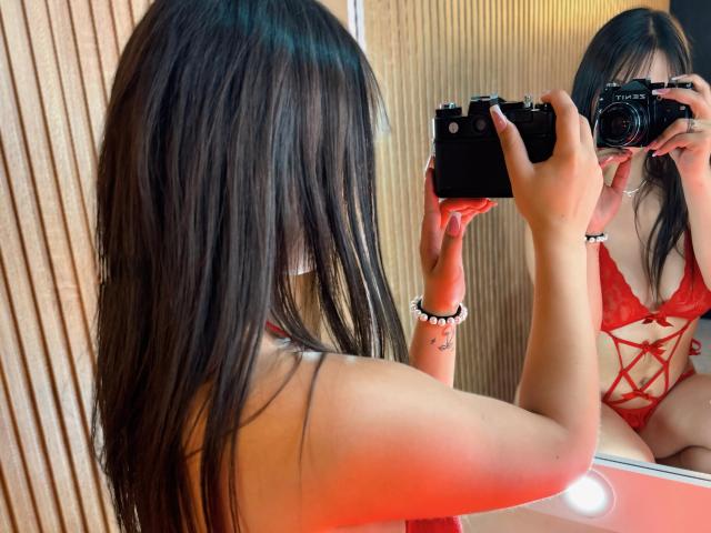 MaddyHot69, com cabelos castanhos longos, vestindo lingerie vermelha de renda, tirando uma selfie em um espelho com uma câmera, mostrando seus seios no reflexo..