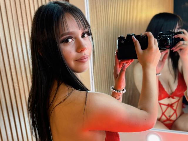 MaddyHot69, com cabelos longos e pretos, vestindo um sutiã de renda vermelho com alças cruzadas, segurando uma câmera, tirando uma selfie, sorrindo, exibindo seus seios no espelho..