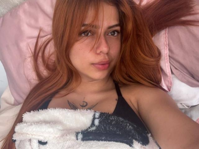 Samanta Carter con lunghi capelli rossi, sdraiata a letto, indossa un top nero, una coperta bianca e soffice, ha un piccolo tatuaggio sul petto, un cuscino rosa e uno bianco, e guarda la fotocamera con un leggero sbuffo..