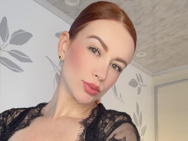 Leahred has red hair, pale skin, and pink lips, wearing a black lace top, with small earrings, and a slight smirk, looking at the camera.