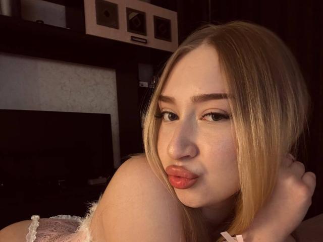 JoetteShigeta with blonde hair, pouty pink lips, and thick eyeliner, looking over her shoulder, showing her bare shoulder and a hint of her white lace top.
