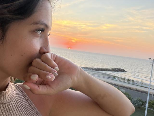 Gabrielafoxxy, la main près de la bouche, montrant un mamelon rose, coucher de soleil sur l'océan, portant un haut beige sans manches, cheveux bruns clairs, yeux verts et vernis à ongles blanc..